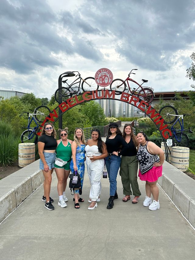Seven friends smiling under a decorative bicycle arch at a brewery courtyard, with industrial tanks and a cloudy sky in the background.