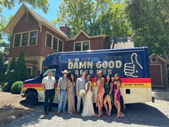 Wedding party in dresses and suits posing beside a blue branded beer bus with a thumbs-up logo on a gravel driveway in front of a red-shingled suburban house on a sunny summer day