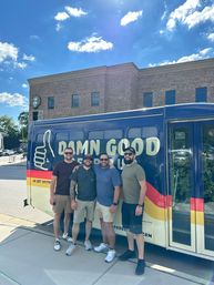 Four friends in casual summer clothes smiling arm‑in‑arm beside a blue and yellow food truck that reads “Damn Good,” parked on a sunny downtown street in front of a brick building and bright blue sky.