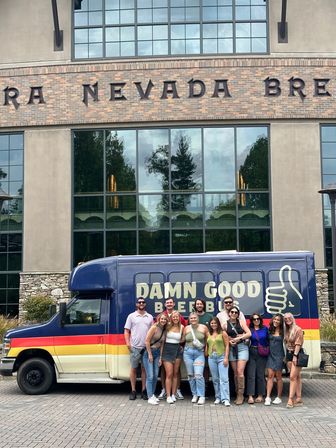 Smiling group of about a dozen adults posing in front of a colorful beer-tour shuttle with a thumbs-up logo parked outside a large brick-and-glass brewery-style building on a daytime outing.
