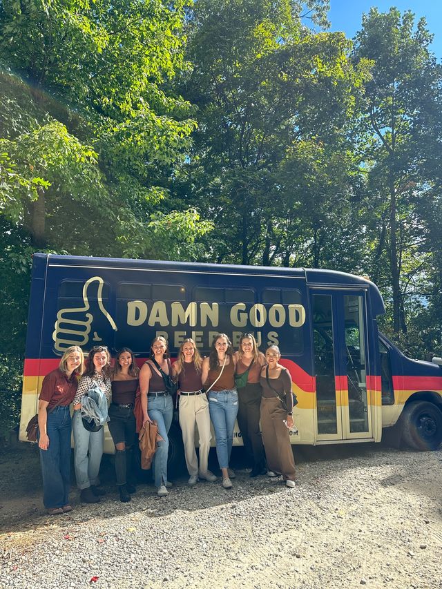 Group of friends smiling and posing in front of a colorful beer shuttle bus parked on a gravel clearing surrounded by sunlit green trees.
