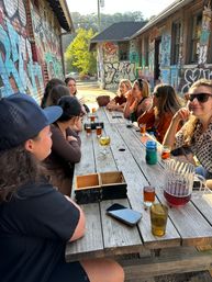 Sunlit urban outdoor patio scene: a group seated at a long wooden picnic table beside graffiti-covered brick walls, sipping beer and drinks from glasses and a pitcher.