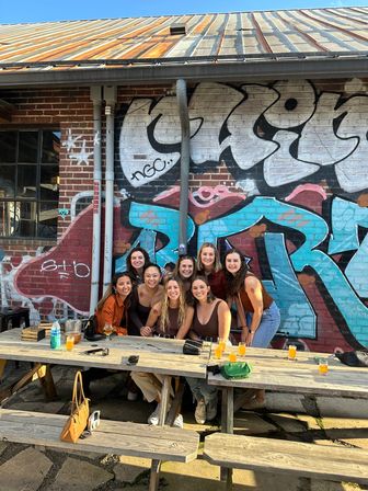 Smiling group of friends gathered at wooden picnic tables on a sunny urban patio in front of a colorful graffiti-covered brick wall, enjoying small beer flights.