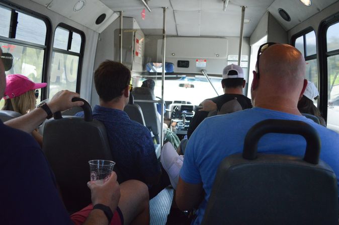 Daytime shuttle bus interior with casually dressed passengers in caps and sunglasses, a hand holding a plastic cup, and a forward view toward the driver — group travel on a minibus.