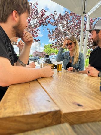 Group of friends chatting and laughing at a wooden outdoor picnic table under a white canopy, sipping small beer glasses on a sunny patio with purple-leaved trees and parked cars in the background.