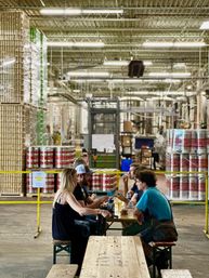 Group of people sampling beers at a long wooden picnic table inside an industrial brewery warehouse, surrounded by stacked kegs, pallets of cans and brewing tanks in the background.