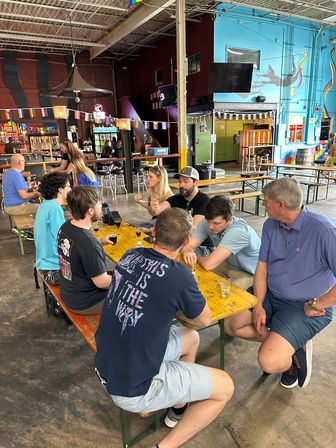 Group of people chatting and tasting beers at yellow picnic tables inside a colorful warehouse-style brewery taproom with murals, string lights, and an industrial ceiling.