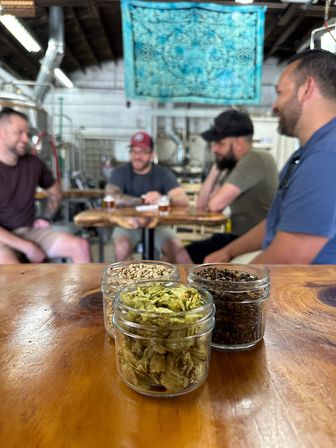 Close-up of three small glass jars holding dried hops and malts on a polished wooden bar, with industrial brewing equipment and a blurred group chatting over beer in a craft brewery taproom.