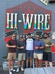 Group of five friends smiling and holding beer glasses on a brewery patio in front of a colorful circus-style mural painted on a brick wall.