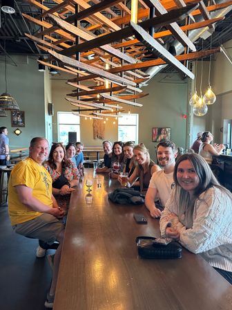 Smiling group of friends at a long wooden communal table in a sunlit craft brewery taproom, holding beers beneath modern wooden ceiling art and hanging bulb lights.