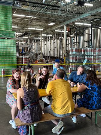 Group of friends laughing and sampling beers at a craft brewery tasting area, seated at a wooden picnic table inside a warehouse with stainless fermentation tanks and stacked cans in the background.