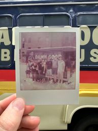 Hand holding a Polaroid of a smiling group posing in front of a colorful vintage bus with navy, red and yellow stripes and bold lettering, outdoor urban scene.