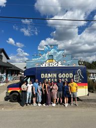 Group of adults posing in front of a colorful beer-tour van parked on a small-town street, with a stepped brick building painted with a sky-and-cloud mural and bright blue, partly cloudy skies overhead.