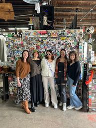 Five friends posing in a rustic industrial taproom with exposed wooden beams, standing in front of a colorful sticker-covered wall; bride-to-be wearing a sash and heart-shaped sunglasses, others in casual fall outfits and boots.