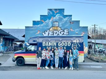 Cheerful group in tropical shirts posing by a retro shuttle-style party van on a street in front of a large blue brick mural of clouds and flying cherubs.