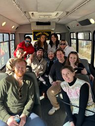 Smiling group of friends inside a sunny shuttle bus, casual winter layers and sunglasses, posing for a daytime road-trip photo with drinks.