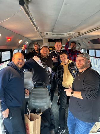 Seven men smiling inside a party bus, wearing casual winter clothes and holding drinks; one man wears a tiara and sash for a playful celebration.