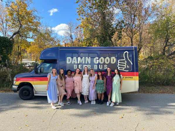 Group of smiling women in pastel dresses posing in front of a blue party bus with a thumbs-up graphic, parked by trees with autumn foliage on a sunny day