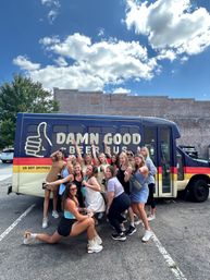 Cheerful group of friends posing with drinks in front of a navy beer tour bus featuring a thumbs-up logo in a downtown parking lot under a bright blue sky with clouds.