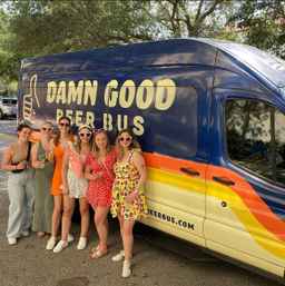 Six friends in summer outfits holding drinks and wearing fun sunglasses, posing in front of a colorful beer-tour van parked under shady oak trees on a sunny street.