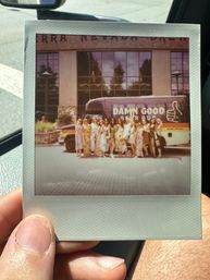 Instant Polaroid held in a car showing a group of women in summer dresses posing in front of a colorful beer tour bus parked outside a brick-and-glass building