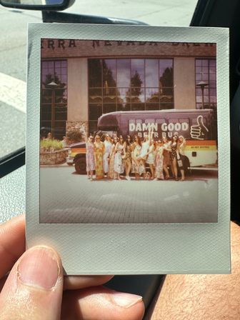 Instant Polaroid held in a car showing a group of women in summer dresses posing in front of a colorful beer tour bus parked outside a brick-and-glass building