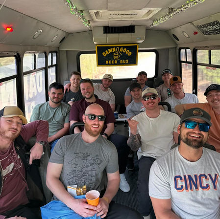 A cheerful group of a dozen men on a beer tour bus, wearing caps and sunglasses and holding cups and snacks beneath a yellow "Damn Good Beer Bus" sign — casual brewery tour vibe (Cincy fanwear visible).