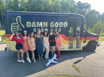 Seven women in colorful retro outfits pose next to a brightly painted beer-themed party bus in a sunny, wooded parking area with an inflatable airplane on the pavement.