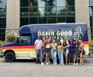 Group of friends smiling and posing in casual summer outfits in front of a colorful “Damn Good Beer Bus” parked outside a stone-and-glass hotel facade, ready for a beer tour.