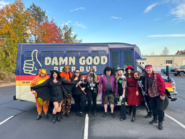 Merry group of adults in Halloween costumes posing in front of a blue-and-yellow party bus parked in a suburban lot with fall foliage and a clear sky.