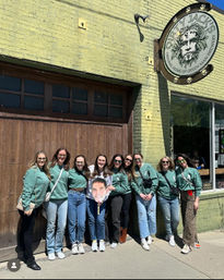 Group photo of ten friends in matching green sweatshirts and jeans posing on a sunny city sidewalk outside a green-brick brewery, one holding a large face cutout.