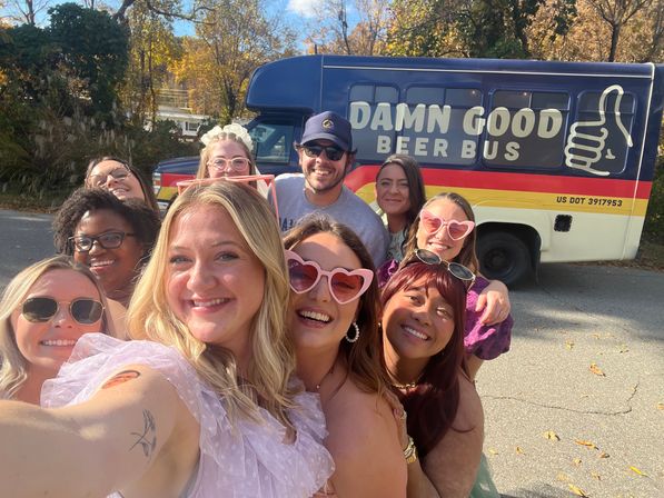 Smiling group selfie of friends wearing sunglasses and heart-shaped shades in front of a blue-and-yellow beer tour bus on a sunny autumn day with fall foliage in the background.