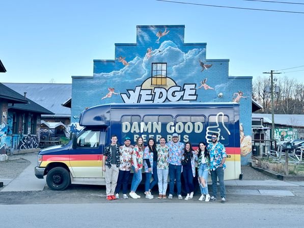 Nine friends in colorful Hawaiian shirts pose by a retro beer shuttle van in front of a stepped blue brick mural of clouds and cherubs on an urban street.