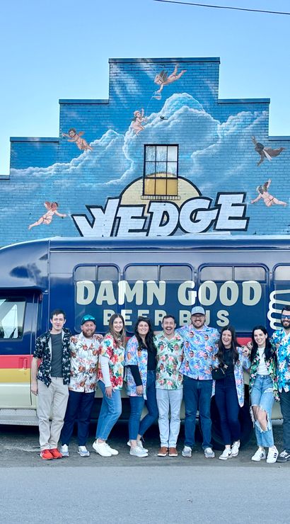 Nine friends in colorful Hawaiian shirts pose by a retro beer shuttle van in front of a stepped blue brick mural of clouds and cherubs on an urban street.