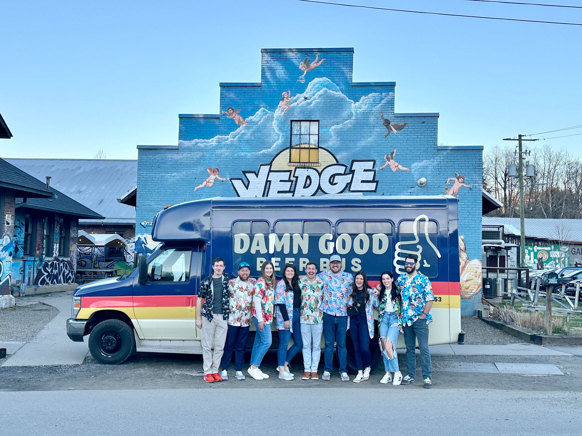 Nine friends in colorful Hawaiian shirts pose by a retro beer shuttle van in front of a stepped blue brick mural of clouds and cherubs on an urban street.