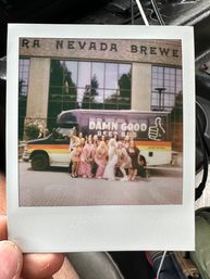 Polaroid photo of a group of friends in colorful dresses posing in front of a retro beer-themed party shuttle outside a Nevada brewery facade.