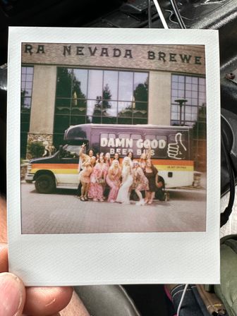 Polaroid photo of a group of friends in colorful dresses posing in front of a retro beer-themed party shuttle outside a Nevada brewery facade.