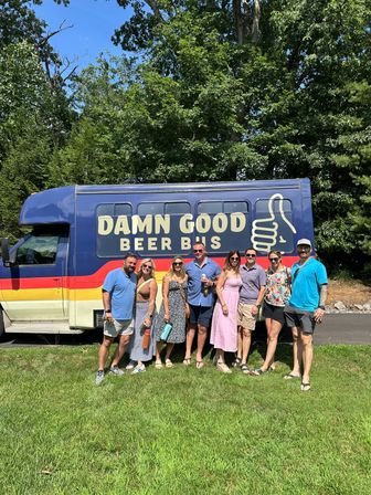 Eight friends smiling and holding drinks in front of a colorful craft beer tour bus with a thumbs-up logo on a sunny summer day in a grassy, tree-lined park