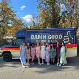 Nine women in pastel and floral dresses smiling and posing in front of a colorful navy-and-yellow party bus with a thumbs-up logo, parked on a tree-lined suburban road with fall foliage and a bright blue sky.