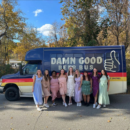 Nine women in pastel and floral dresses smiling and posing in front of a colorful navy-and-yellow party bus with a thumbs-up logo, parked on a tree-lined suburban road with fall foliage and a bright blue sky.