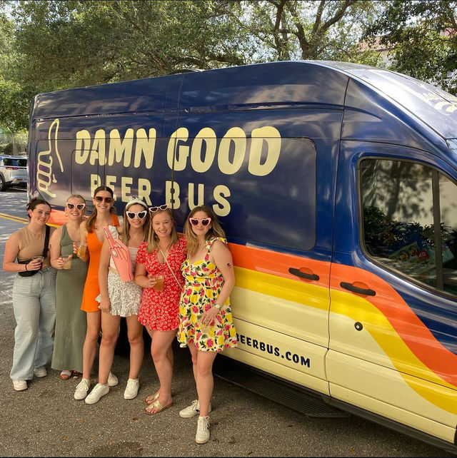 Six women in colorful summer dresses and heart-shaped sunglasses holding drinks, posing beside a navy-and-cream beer bus van with a rainbow stripe parked on a tree-lined street