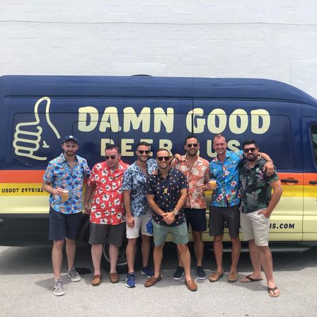 Seven men in bright Hawaiian shirts smiling and holding drinks while posing in front of a colorful blue-and-yellow van with a large thumbs-up logo, parked outdoors on a sunny day.