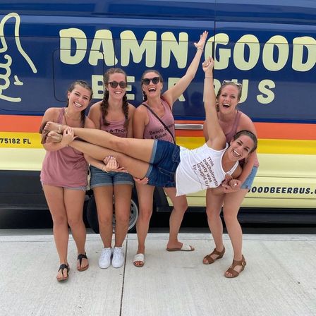 Five smiling friends on a sunny sidewalk, four women holding the fifth horizontally in front of a colorful blue-and-yellow party van, playful bachelorette celebration vibe.