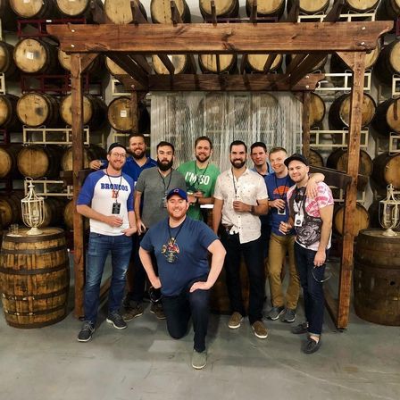 Group of friends posing under a wooden pergola in a craft brewery barrel room with stacked oak barrels, smiling and holding beers.