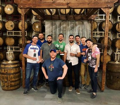 Group of friends posing under a wooden pergola in a craft brewery barrel room with stacked oak barrels, smiling and holding beers.