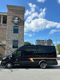 Black shuttle van with retro rainbow stripe parked beside a brick-and-stone commercial building featuring a carved leafy face on the wall, bright blue sky with puffy clouds and midrise apartments in the background