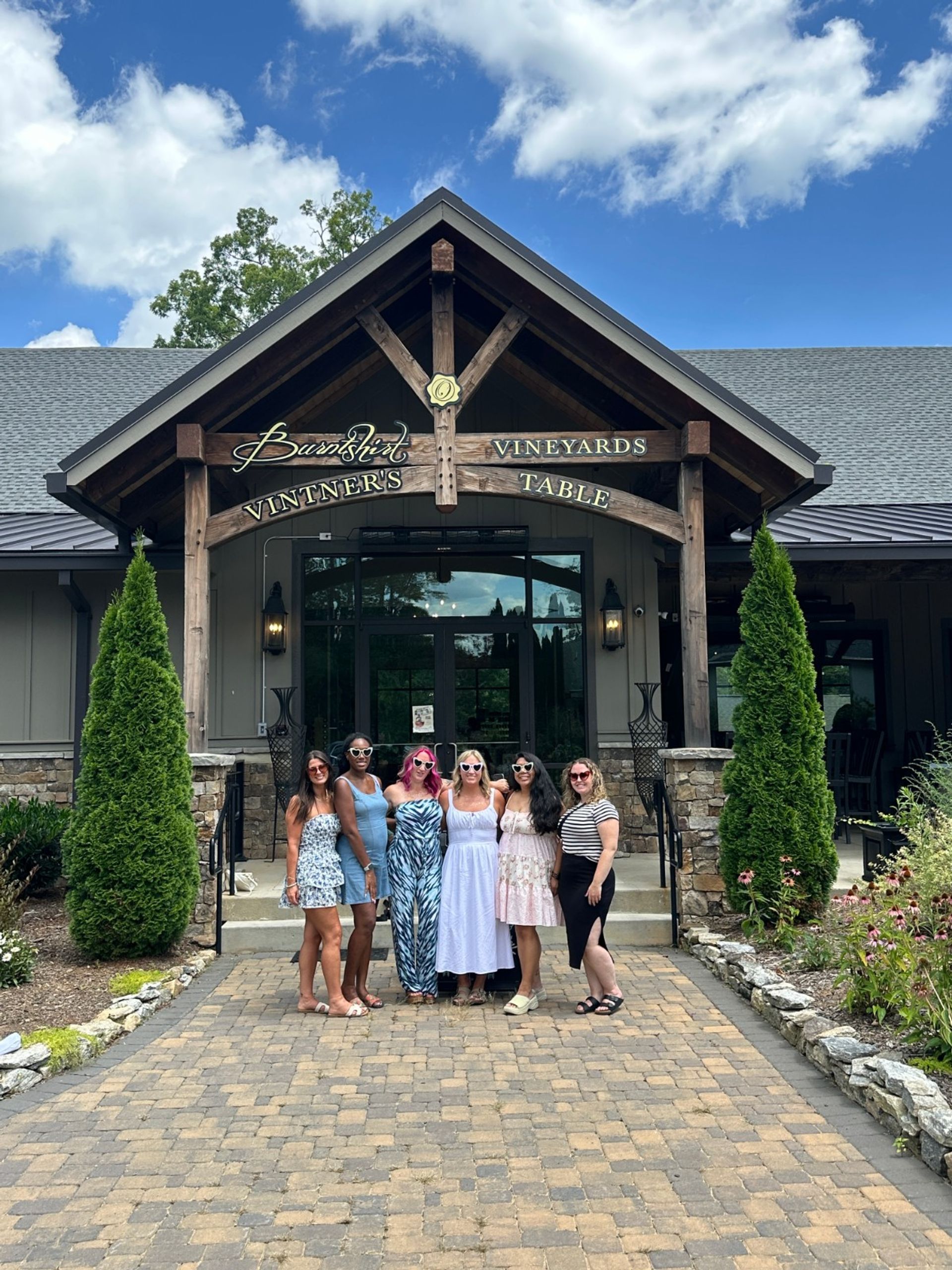 Six women in summer dresses and sunglasses pose on a cobblestone path in front of a winery tasting-room entrance with a wooden gabled porch, stone planters, evergreen shrubs and a bright blue sky.