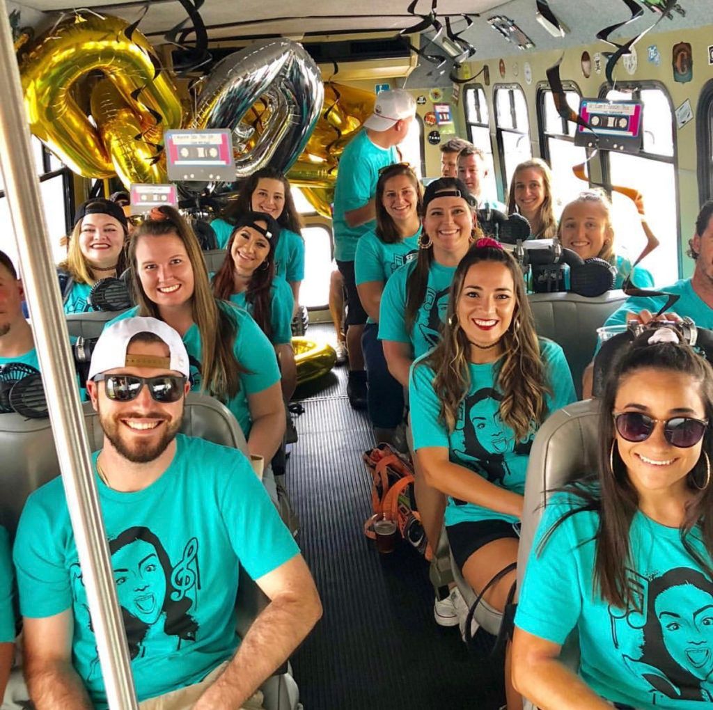 Smiling group in matching teal shirts celebrating on a decorated party bus with gold and silver balloons and retro cassette decorations.