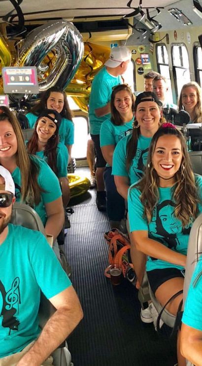Smiling group in matching teal shirts celebrating on a decorated party bus with gold and silver balloons and retro cassette decorations.