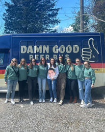 Nine friends in matching green sweatshirts smiling and posing in front of a blue-and-red beer tour bus with a large thumbs-up logo, holding an oversized face cutout on a sunny day in a gravel lot.